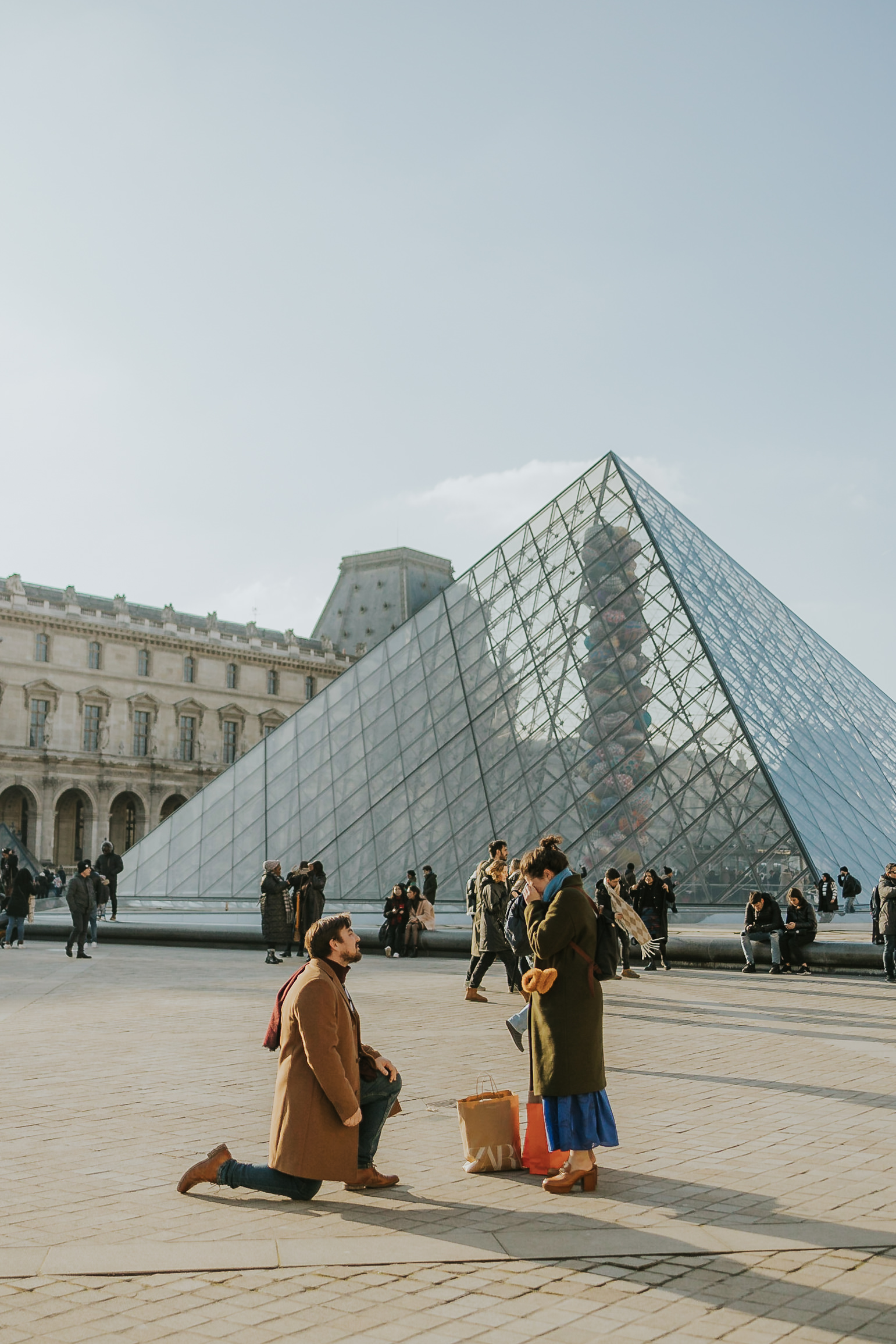 Man kneeling to propose in front of the Louvre Pyramid during a surprise Paris engagement