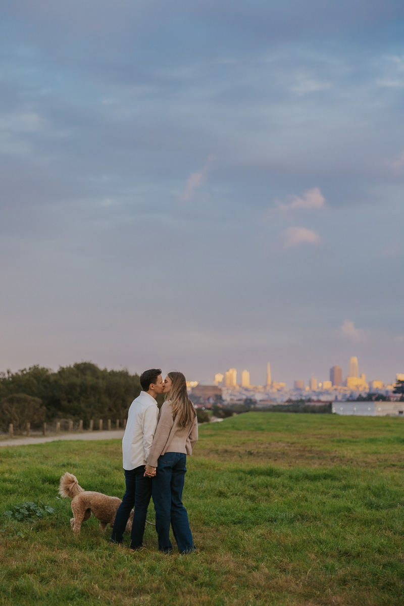 Couple holding hands kissing while San Francisco glows in the the sunset light in the background